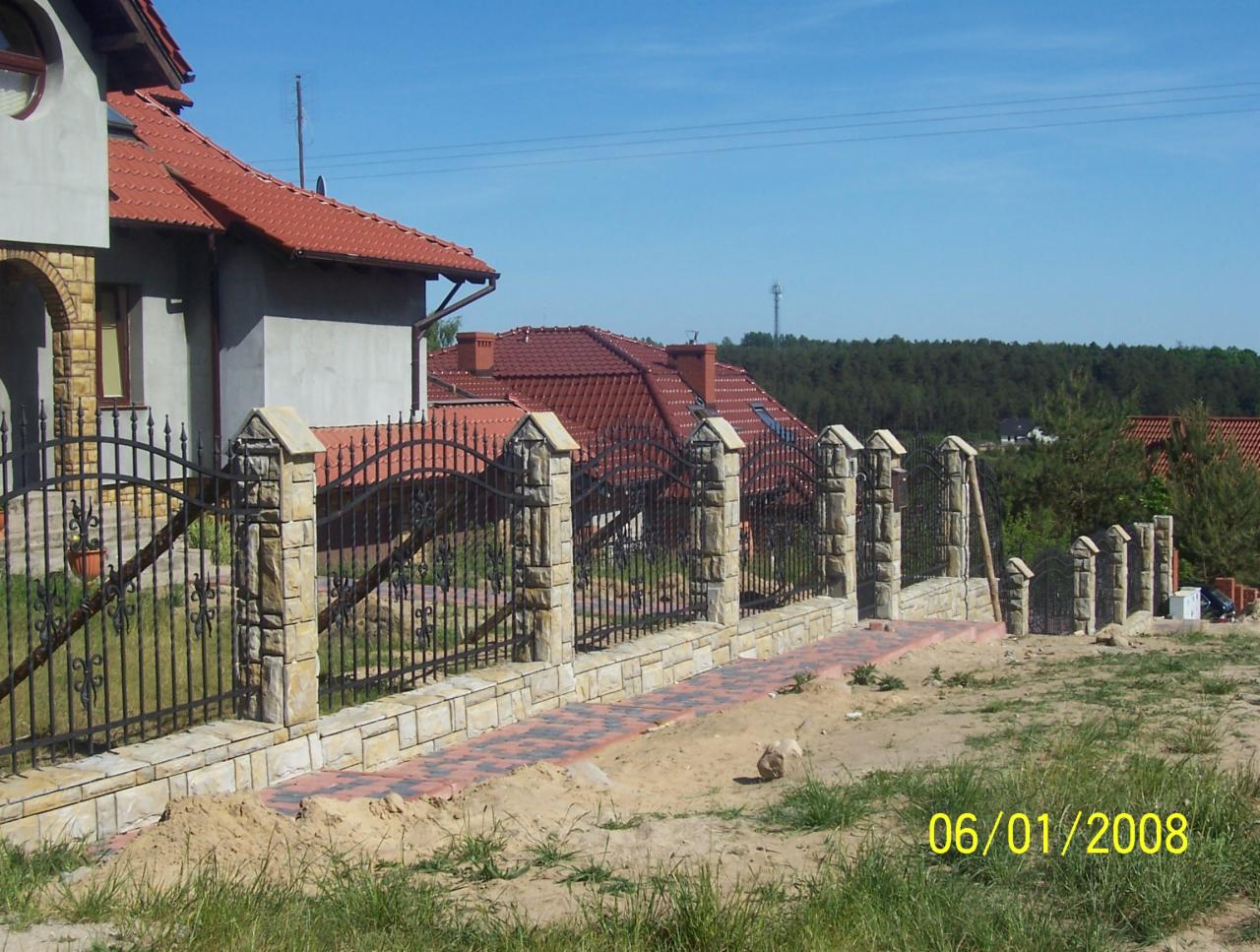 Nowoczesne ogrodzenie z kutymi elementami i kamiennymi słupkami, świeżo po montażu. Widoczny fragment domu z czerwoną dachówką i ścieżka z kostki.