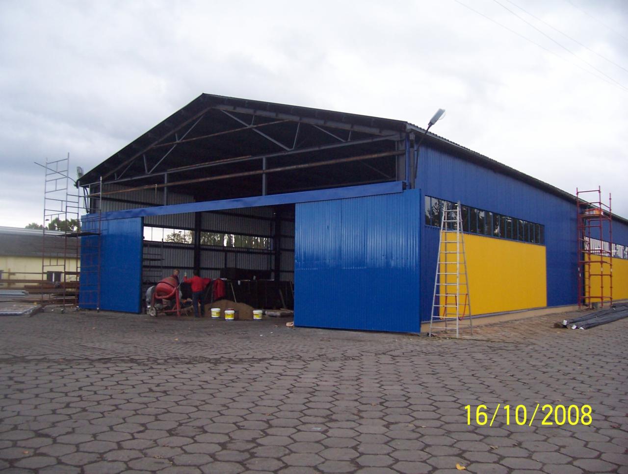 Niebieski budynek magazynowy z żółtym pasem, częściowo otwarte przesuwne drzwi, rusztowania i drabina, widoczna betoniarka oraz data 16/10/2008 na brukowanym placu.