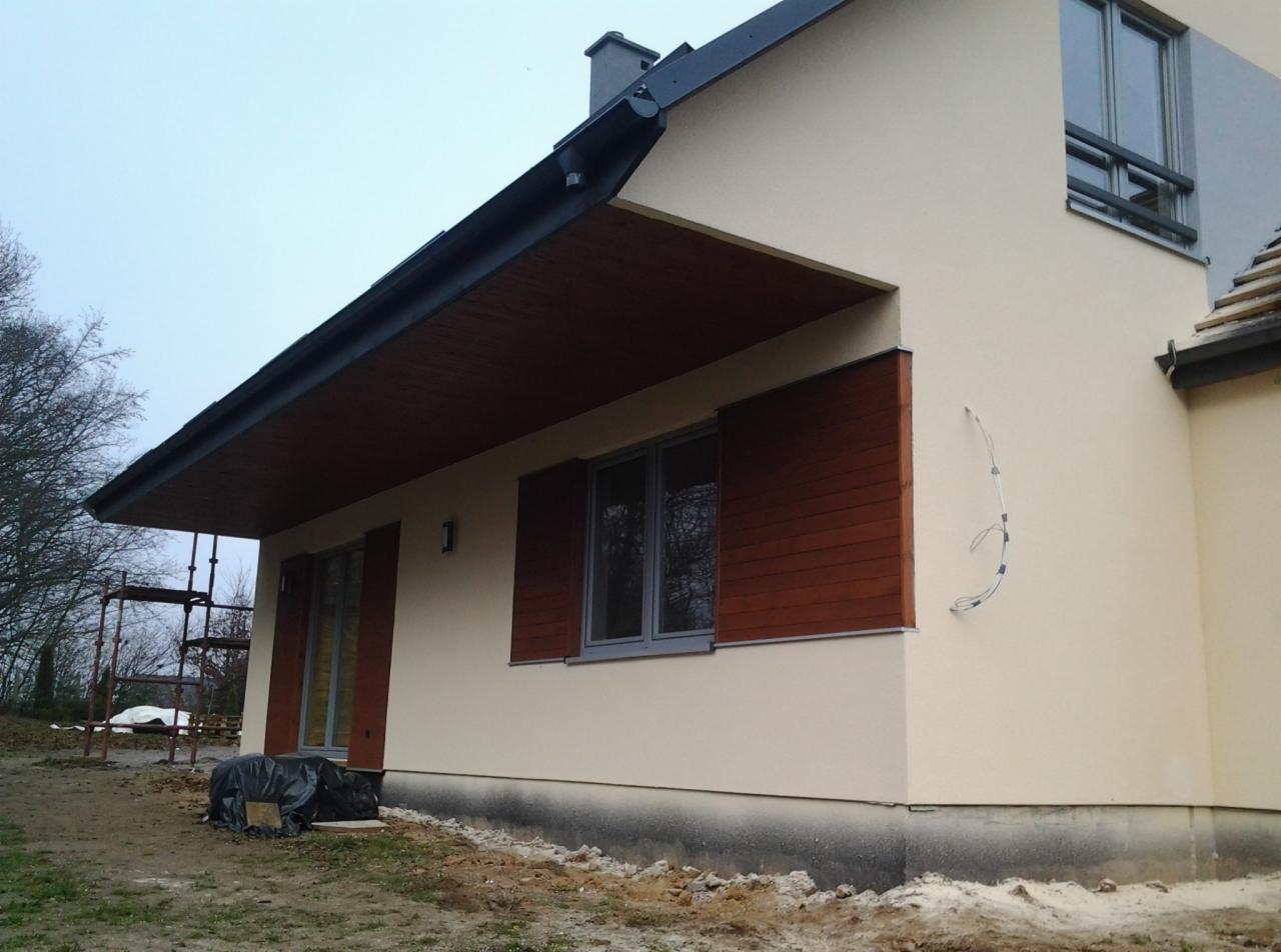 Elewacja domu jednorodzinnego z drewnianymi elementami, świeżo po tynkowaniu. Widoczne rusztowanie i kabel wystający ze ściany. Kremowa kolorystyka budynku.