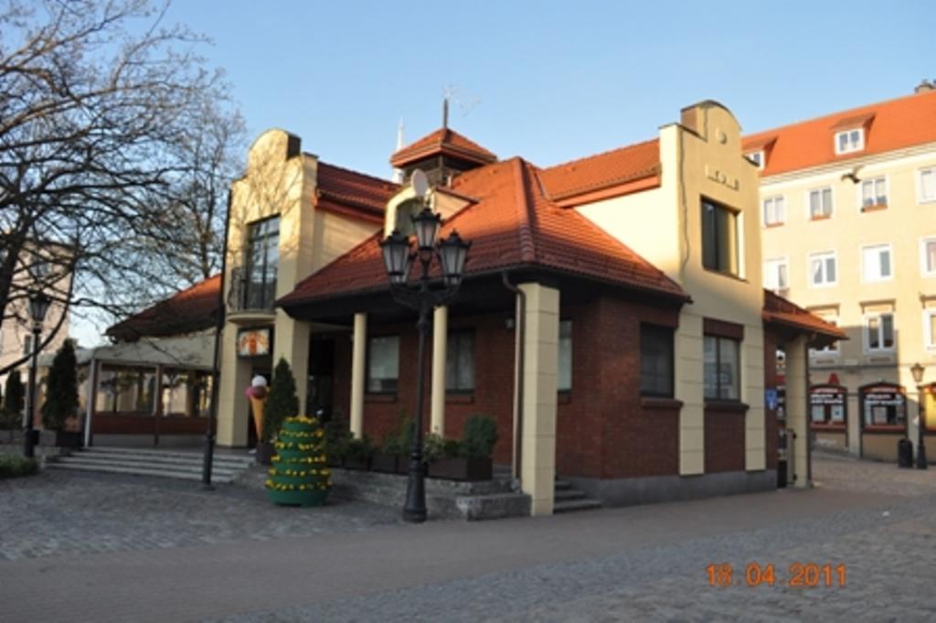 Dwupiętrowy budynek z czerwonej cegły i beżowego tynku, z czerwoną dachówką, oświetlony wczesnym słońcem. Na pierwszym planie latarnia i dekoracje.