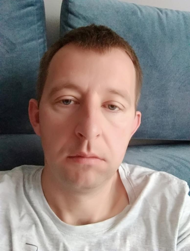 Portret mężczyzny z krótkimi, jasnymi włosami na tle niebieskiej sofy. Ubrany w szarą koszulkę. Ujęcie od przodu, z bliska, z neutralnym wyrazem twarzy.