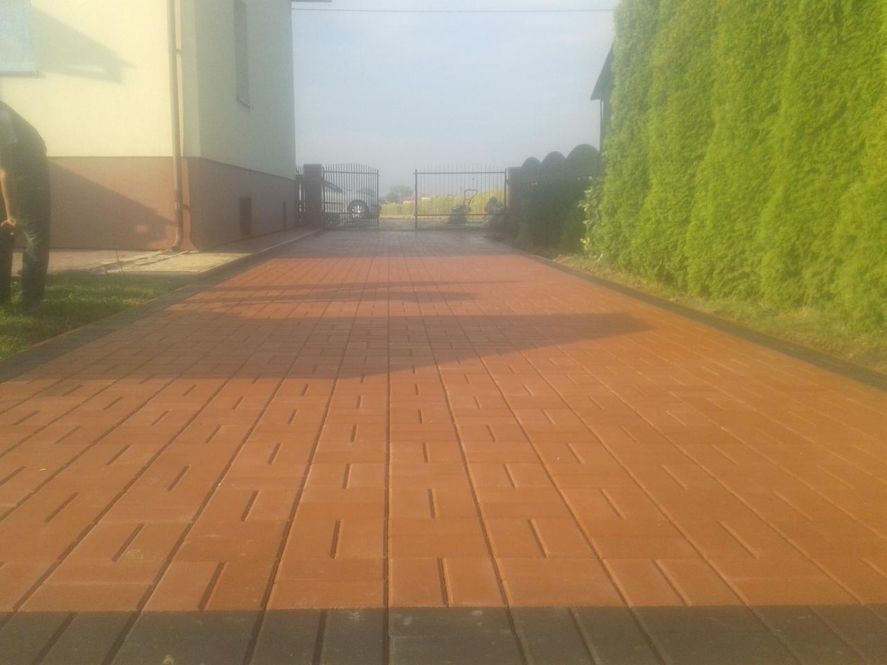 Ułożona, brązowa kostka brukowa z ciemniejszym obramowaniem tworzy wzór na podjeździe. W tle brama, samochód i żywopłot. Częściowo widoczna elewacja budynku.