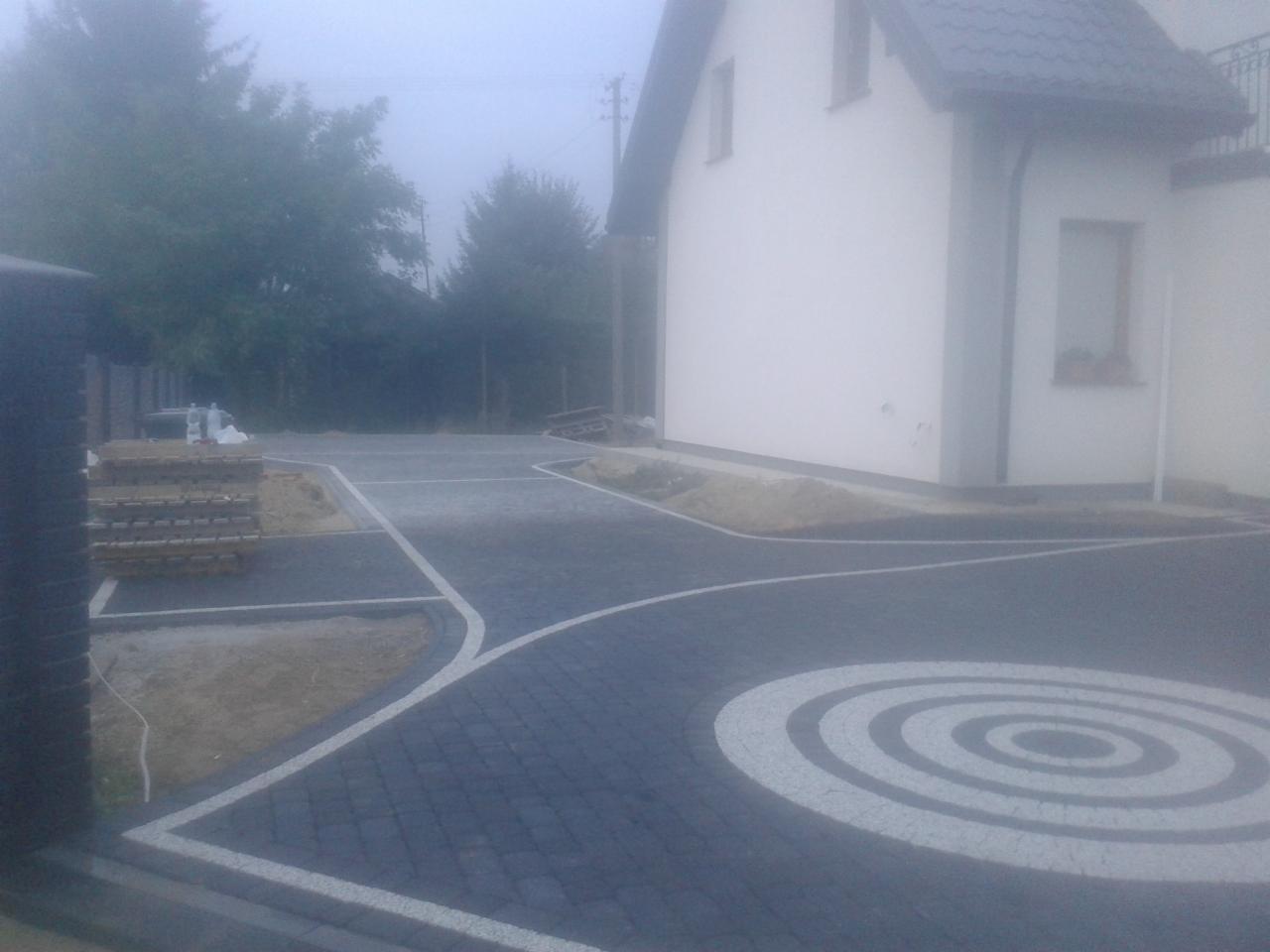 Nowoczesna nawierzchnia z kostki brukowej w odcieniach szarości z białym wzorem koła na podjeździe, częściowo ułożona, z paletą kostki w tle i fragmentem budynku.