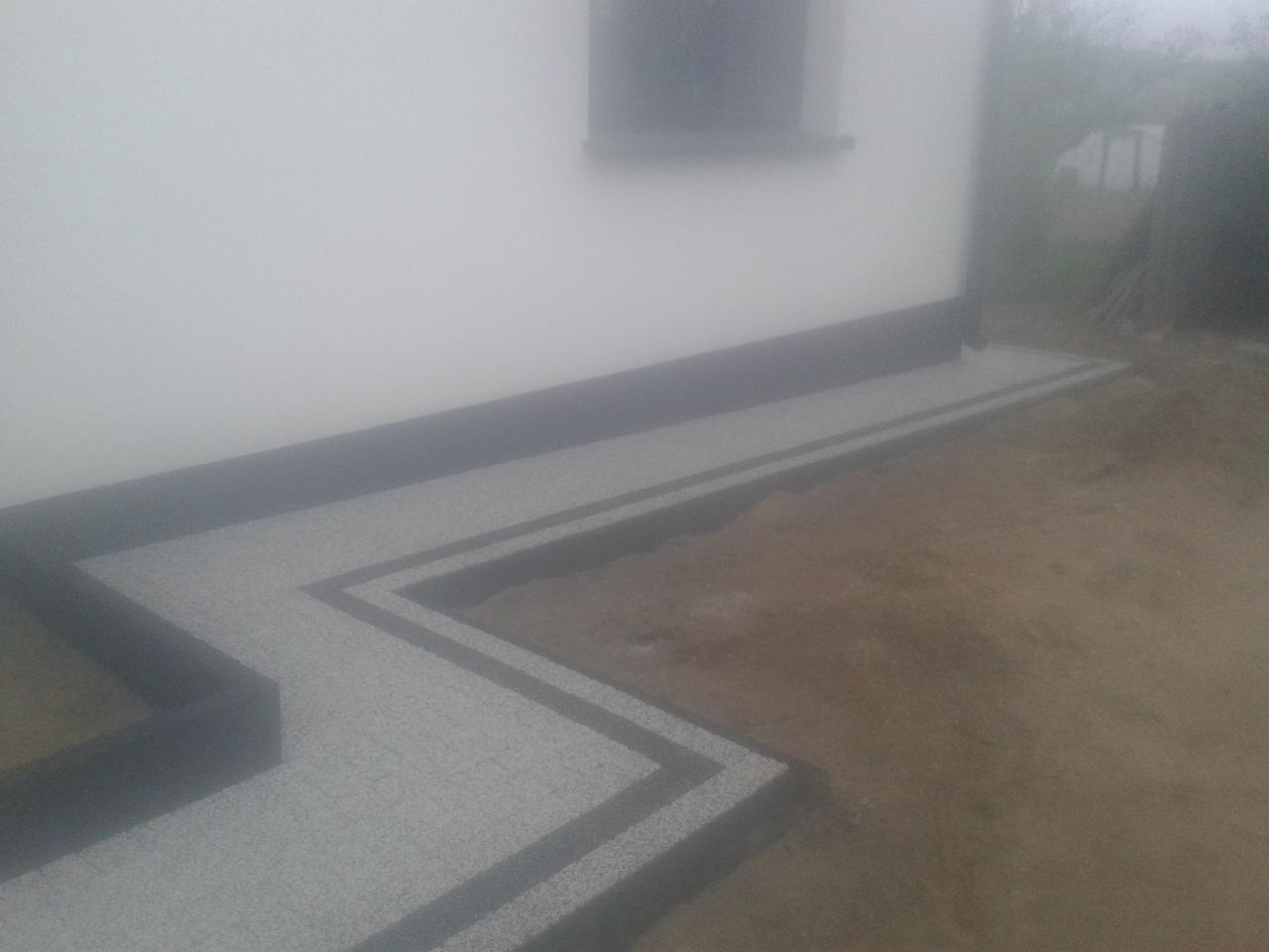 Świeżo ułożony chodnik z jasnoszarej kostki z ciemnoszarą obwódką przy białej ścianie budynku, widoczny fragment okna i ziemia.