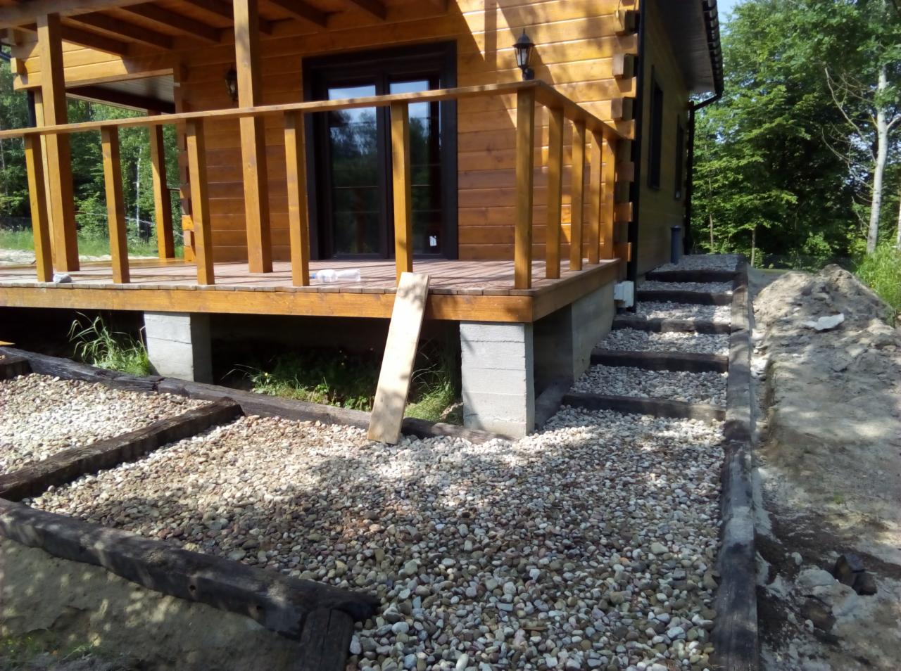 Drewniany dom z tarasem i schodami, otoczony żwirową ścieżką z drewnianymi obrzeżami, częściowo ułożoną, z widocznym fragmentem niezagospodarowanego terenu.