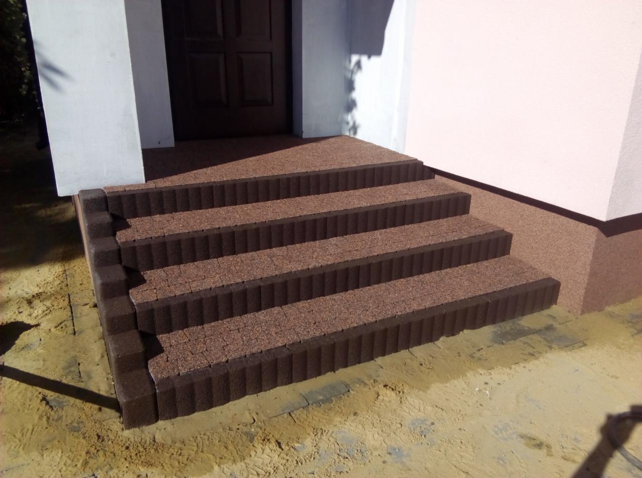 Czterostopniowe schody wejściowe wyłożone brązową kostką brukową o nieregularnej fakturze, z ciemnobrązowymi obrzeżami i fugami, prowadzące do ciemnych drzwi wejściowych w jasnym budynku,...