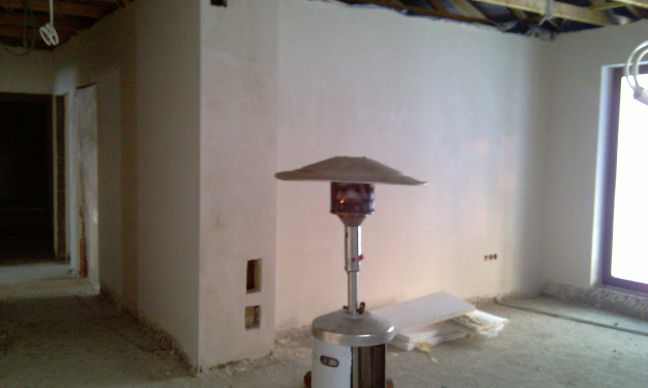 Wnętrze w trakcie tynkowania maszynowego z widocznym nagrzewnicą gazową, surowym stropem i przygotowanymi otworami pod instalacje.