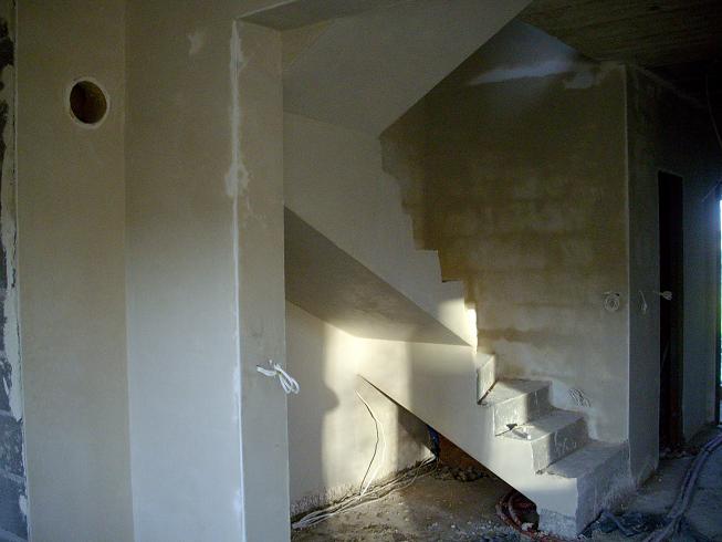 Wnętrze w stanie surowym po tynkowaniu maszynowym, widoczne schody wylewane z betonu, ściany z widocznymi przewodami elektrycznymi i otworem pod gniazdko.