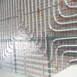 Białe rurki ogrzewania podłogowego zamocowane do ściany z folią w kratkę, widoczne czarne uchwyty mocujące i fragment podłogi z wystającymi końcówkami rurek.