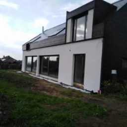 Nowoczesny dom z białą elewacją i grafitową dachówką, duże okna, widok z zewnątrz na trawnik w trakcie prac wykończeniowych.