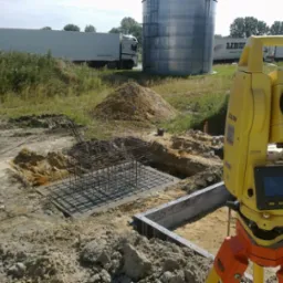 Żółty teodolit geodezyjny na statywie obok wykopu zbrojonego fundamentu pod budowę, w tle zbiornik i ciężarówki. Widok na plac budowy w słoneczny dzień.