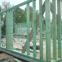 Wznoszenie konstrukcji ścian budynku z impregnowanego drewna, widoczne pionowe słupki i fragment fundamentu, w tle zieleń drzew i biały samochód dostawczy.