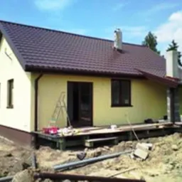 Żółty dom w trakcie budowy z brązowym dachem, widoczne rusztowanie, niezabezpieczony teren i materiały budowlane na zewnątrz.