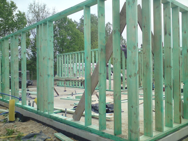 Wznoszenie konstrukcji ścian budynku z impregnowanego drewna, widoczne pionowe słupki i fragment fundamentu, w tle zieleń drzew i biały samochód dostawczy.