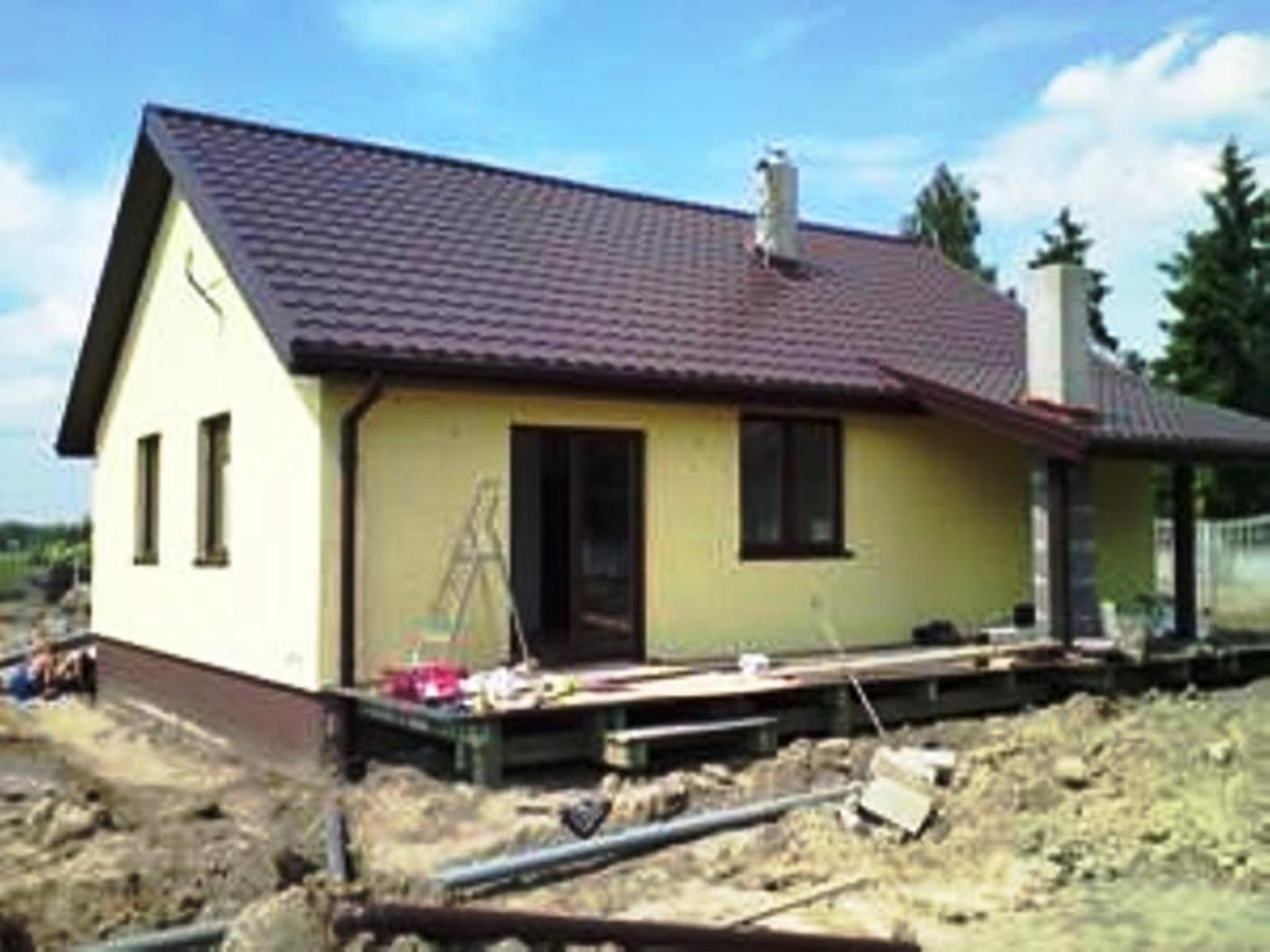 Żółty dom w trakcie budowy z brązowym dachem, widoczne rusztowanie, niezabezpieczony teren i materiały budowlane na zewnątrz.