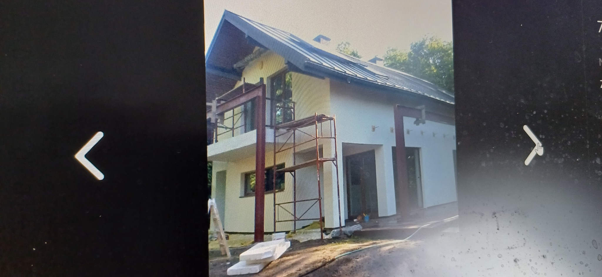 Dwupiętrowy dom w trakcie prac elewacyjnych, widoczne rusztowania, częściowo otynkowana fasada i metalowe elementy konstrukcyjne.