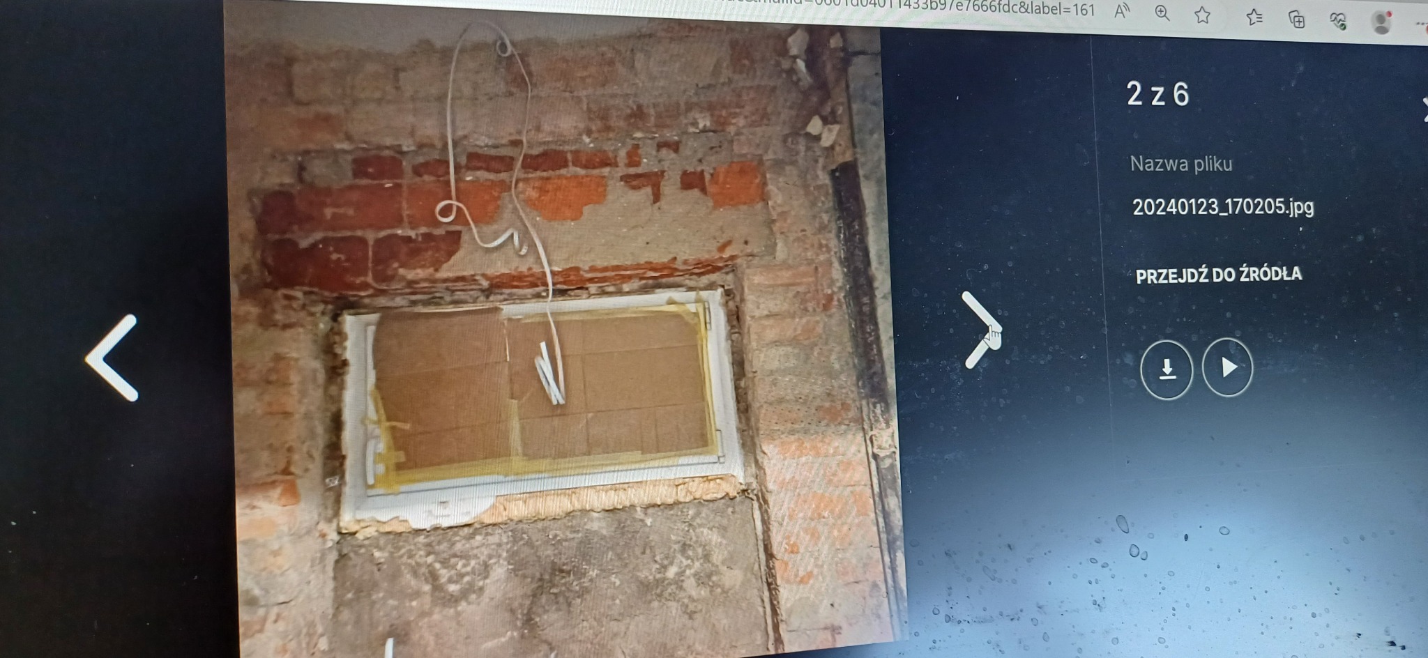 Stara ceglana ściana z częściowo usuniętym tynkiem, widoczne przygotowania do montażu okna, zabezpieczonego kartonem i taśmą malarską, zwisający kabel elektryczny.