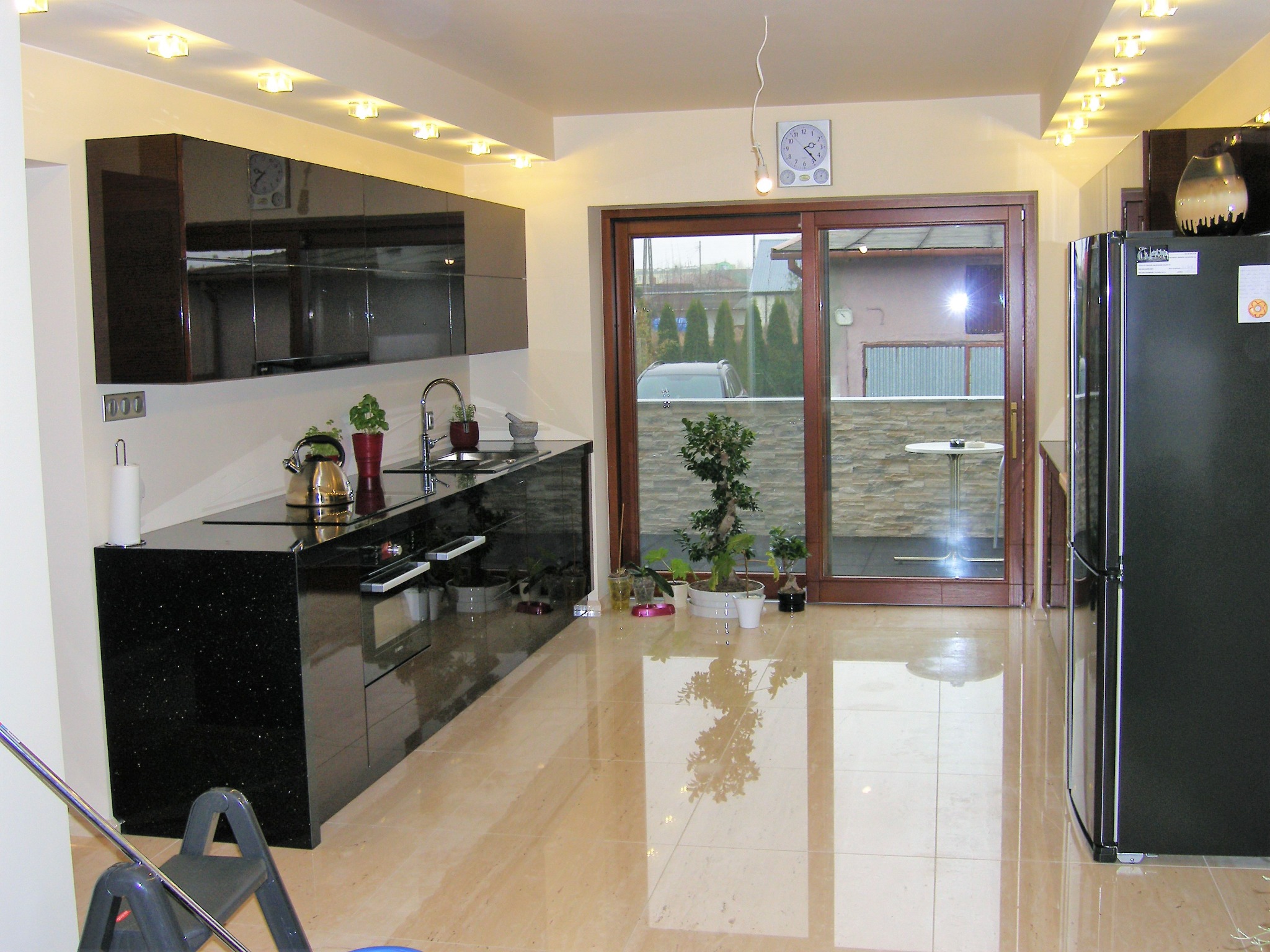 Nowoczesna kuchnia z błyszczącą czarną zabudową, marmurową podłogą i przesuwnymi drzwiami balkonowymi z brązową ramą, przez które widać ogród z tujami.