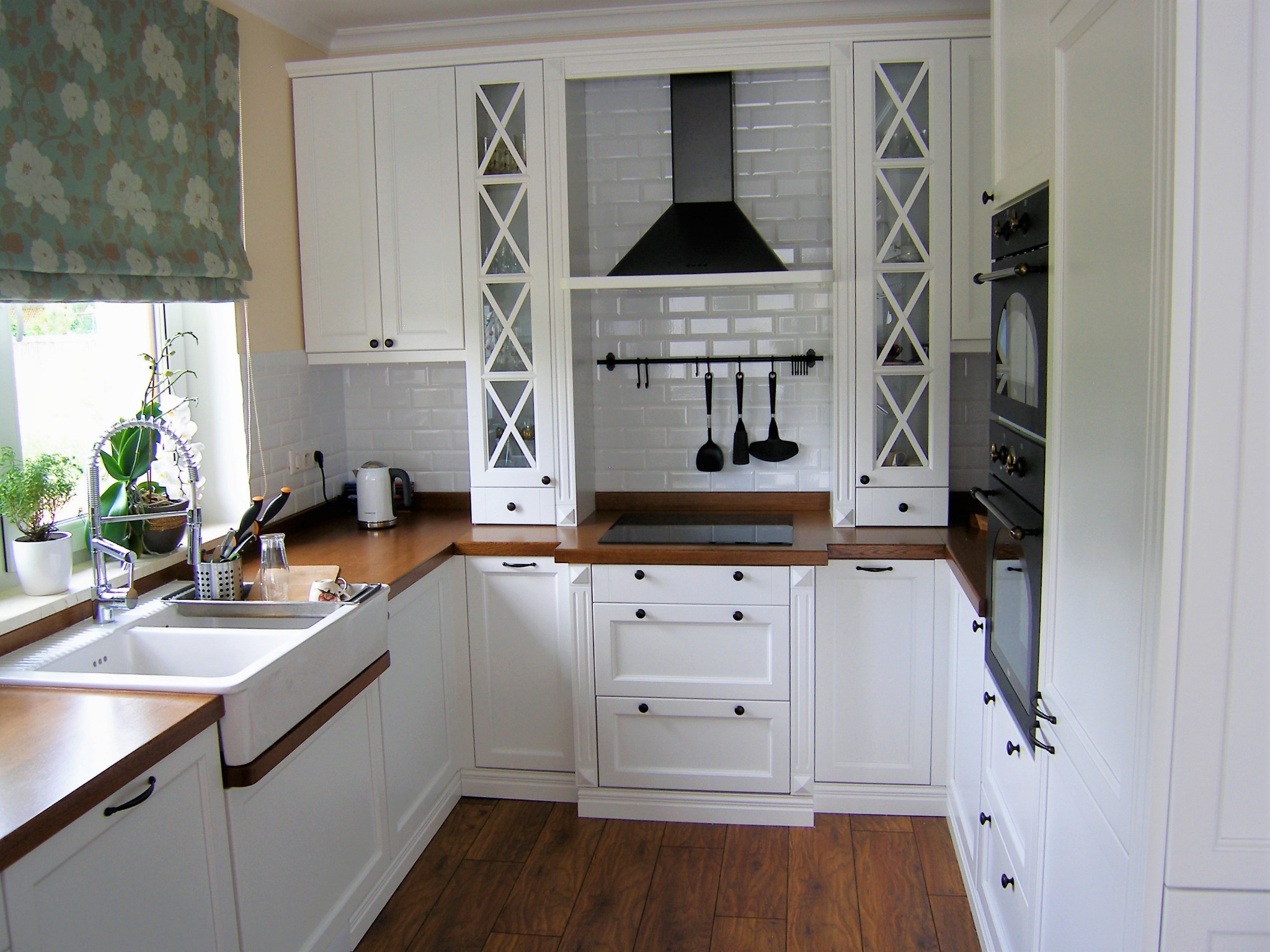 Jasna kuchnia w stylu rustykalnym z białymi szafkami, drewnianym blatem i podłogą, widok na okno z roletą w kwiaty i zlewozmywak farmerski