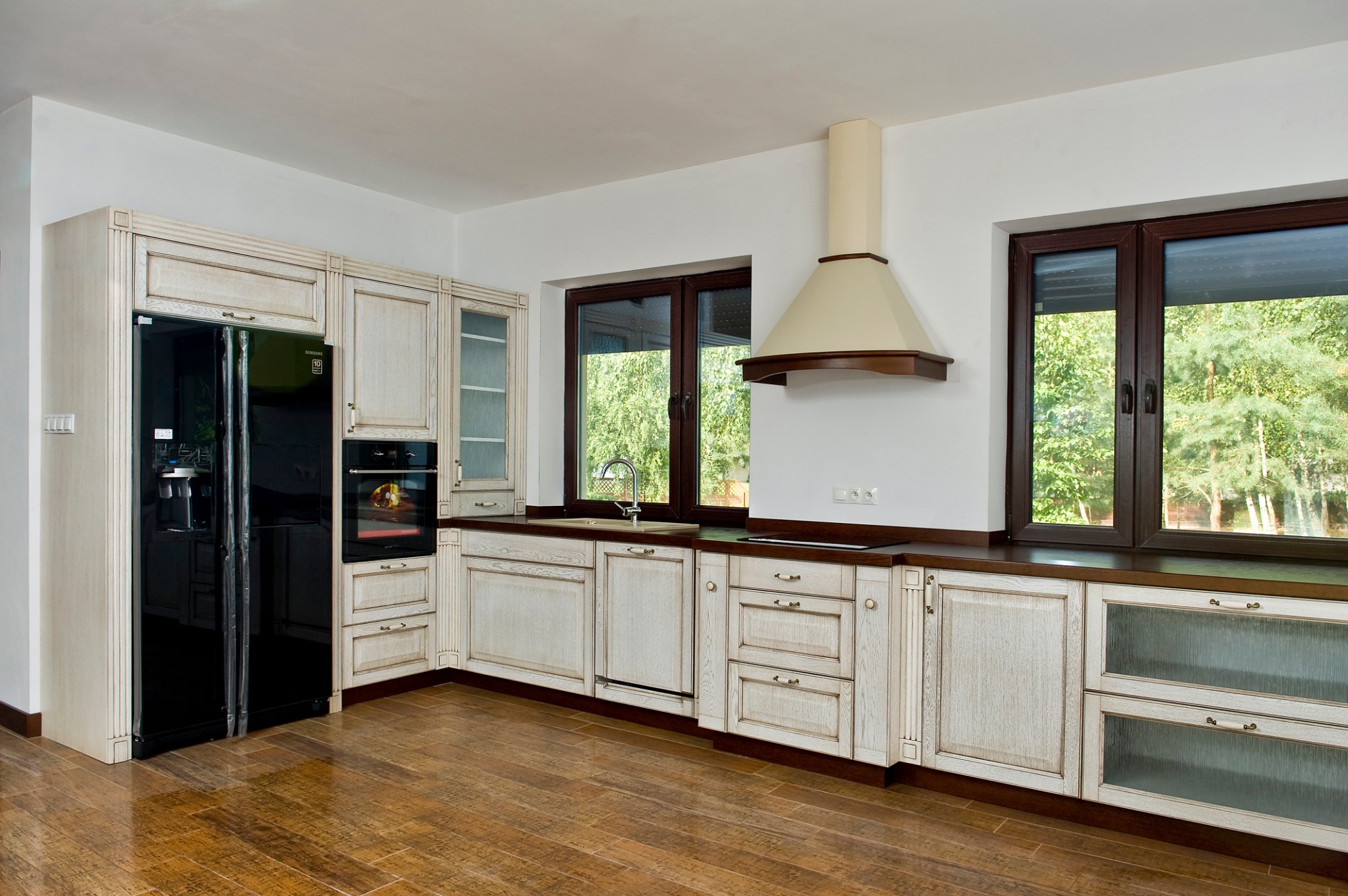 Jasna kuchnia w stylu rustykalnym z drewnianą podłogą, ciemnymi ramami okiennymi i czarną lodówką z dystrybutorem wody.