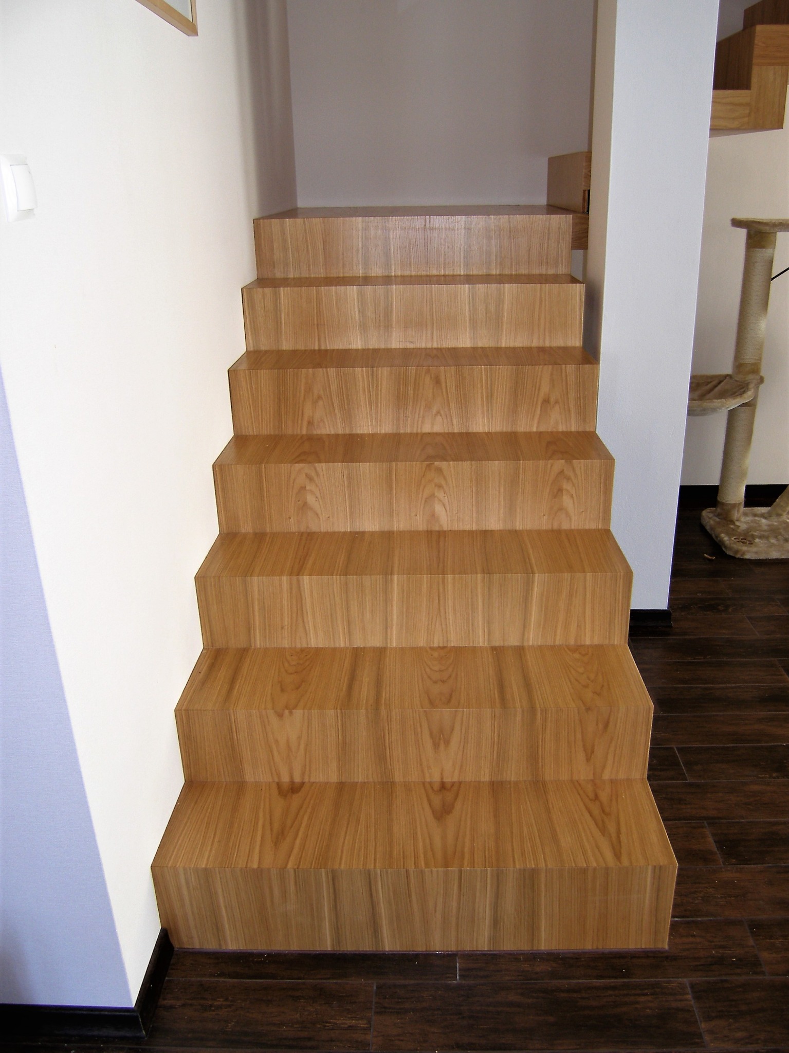 Schody wewnętrzne wykończone drewnopodobną okładziną, z widoczną strukturą drewna, prowadzące do góry w jasnym wnętrzu z ciemną podłogą.