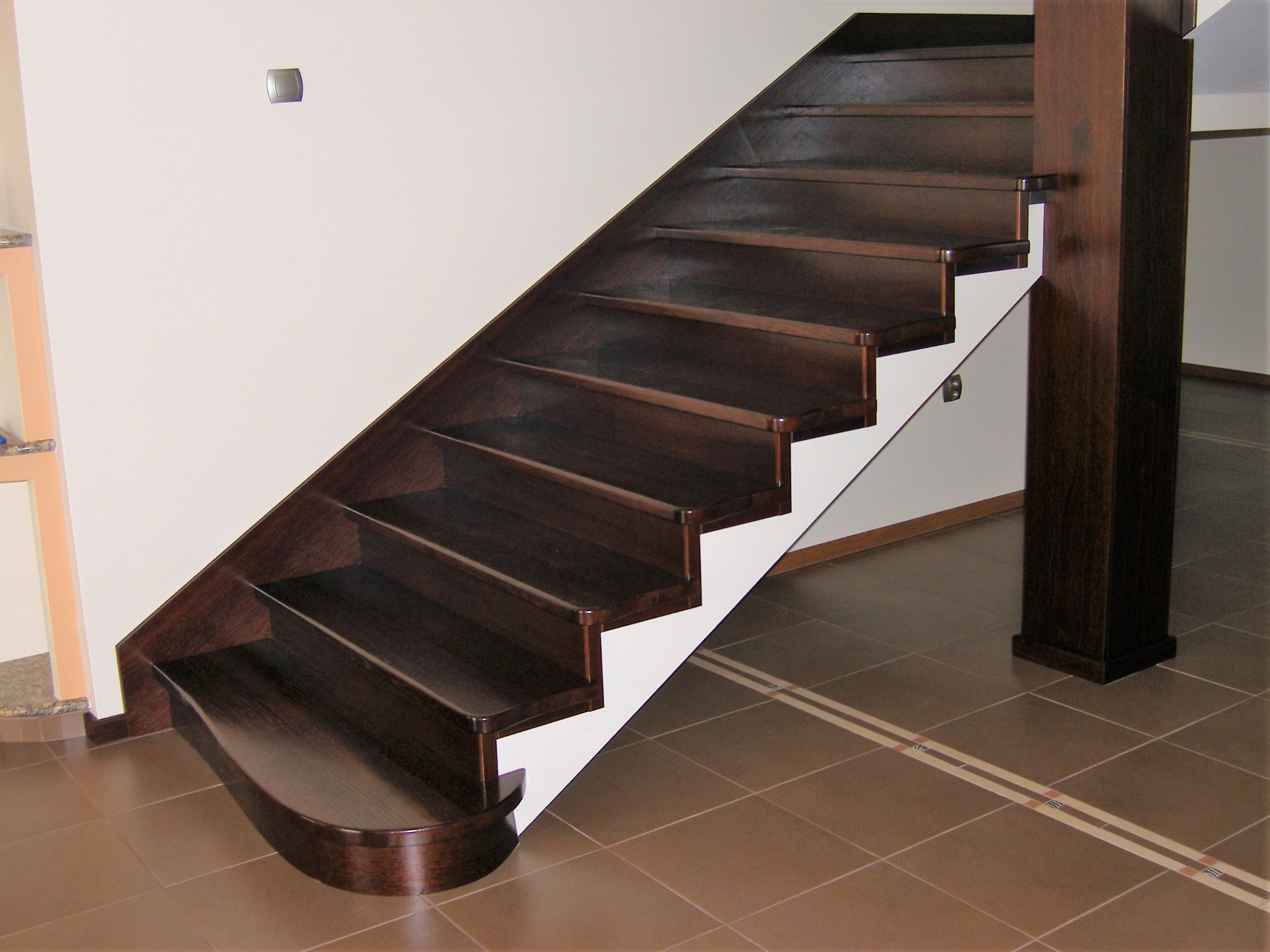Schody drewniane o ciemnym odcieniu z białymi podstopnicami, widoczne z boku, z zaokrąglonym pierwszym stopniem i drewnianym słupem nośnym, na tle brązowej podłogi z płytek.