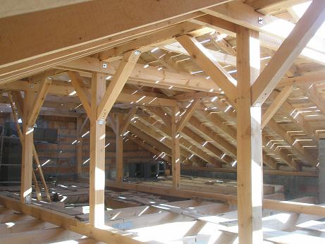 Wnętrze domu w trakcie budowy, widoczne drewniane elementy konstrukcyjne dachu i ścian, surowy stan wykończenia z widoczną cegłą i pustakami.