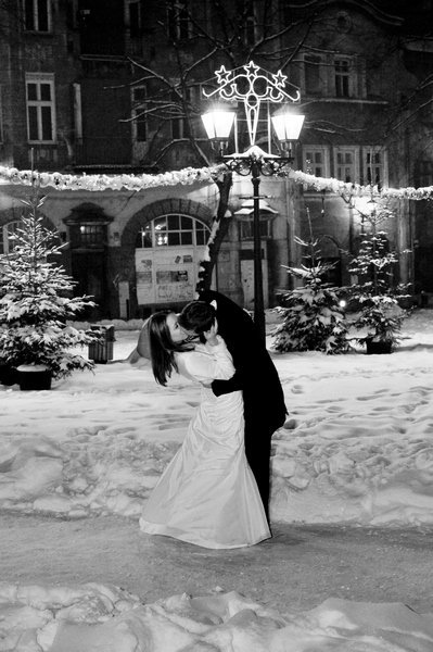 Czarno-białe zdjęcie całującej się pary młodej na tle zaśnieżonego placu miejskiego z oświetloną latarnią ozdobioną gwiazdami i choinkami.