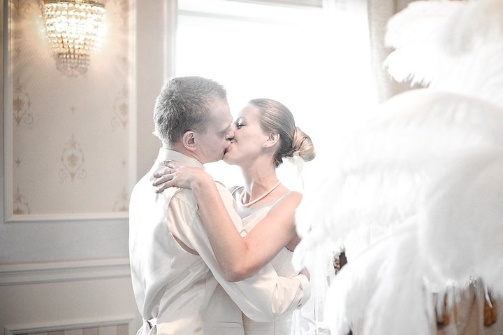 Para młoda w objęciach podczas pocałunku, kobieta w perłowym naszyjniku i upiętych włosach, mężczyzna w kamizelce, dekoracje z piór i kryształowy żyrandol w tle.