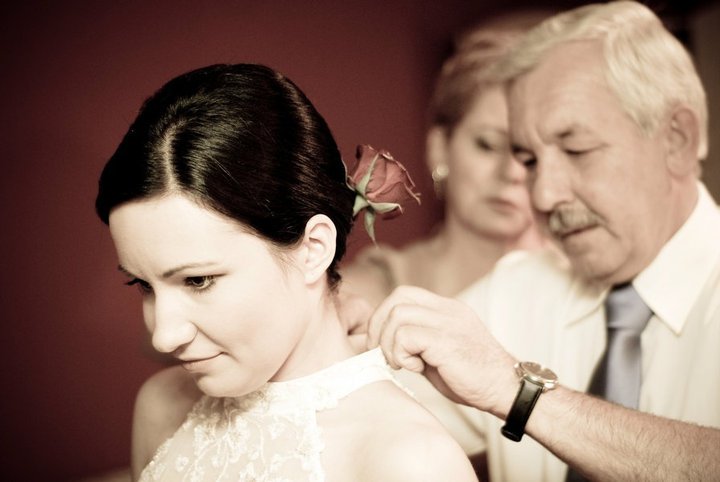 Panna młoda z różą we włosach, dopinana przez mężczyznę w koszuli z krawatem, w tle kobieta