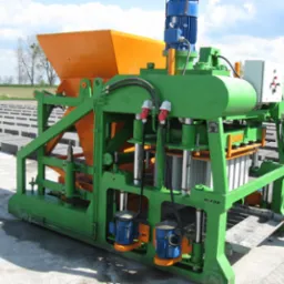 Maszyna do produkcji betonowych bloczków z pomarańczowym zasobnikiem i zieloną ramą, na betonowej powierzchni z ułożonymi bloczkami w tle.