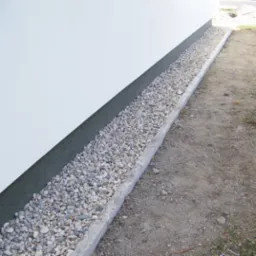 Wykończona elewacja budynku z szarą listwą cokołową i drenażem opaskowym z jasnych kamyków, oddzielonym betonową obwódką od ziemi.