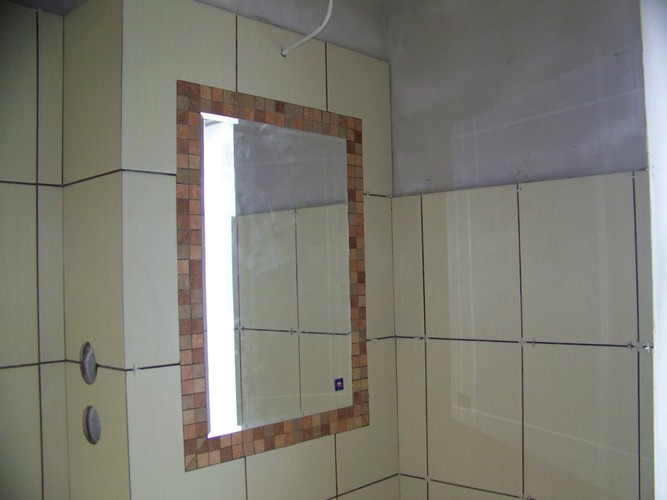 Ściana w trakcie kafelkowania jasnymi płytkami, lustro z mozaikową ramą, widoczne krzyżyki dystansowe i otwory na rury.