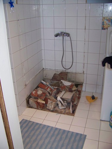 Prysznic w trakcie demontażu: zdemolowana podłoga, gruz ceglany i resztki płytek w miejscu brodzika, widoczna bateria prysznicowa i fragment niebieskiego dywanika.