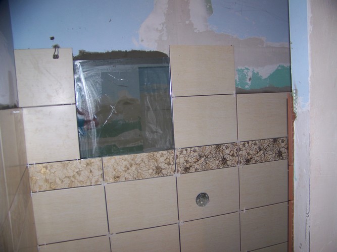 Proces kafelkowania ściany w łazience z użyciem beżowych płytek z dekoracyjnym pasem w kwiaty, widoczne krzyżyki dystansowe i otwór na instalację elektryczną.