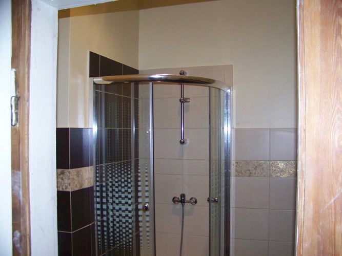 Wykończona kabina prysznicowa z chromowanymi elementami, widoczna przez częściowo otwarte drewniane drzwi. Ściany wyłożone beżowymi i brązowymi płytkami z dekoracyjnym pasem. Część kabiny ma wzór...