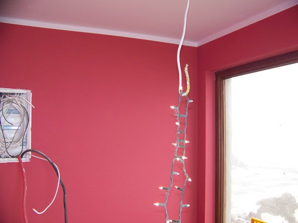 Pokój z pomalowanymi na różowo ścianami, widoczne kable elektryczne w puszce podtynkowej, zwisające lampki choinkowe i okno z widokiem na zewnątrz.