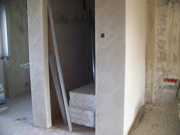 Wnętrze w trakcie remontu w Strzelinie: surowe ściany z widocznymi instalacjami elektrycznymi, fragment otynkowanej ściany z gniazdkiem elektrycznym, styropian i profile aluminiowe oparty o ścianę,...