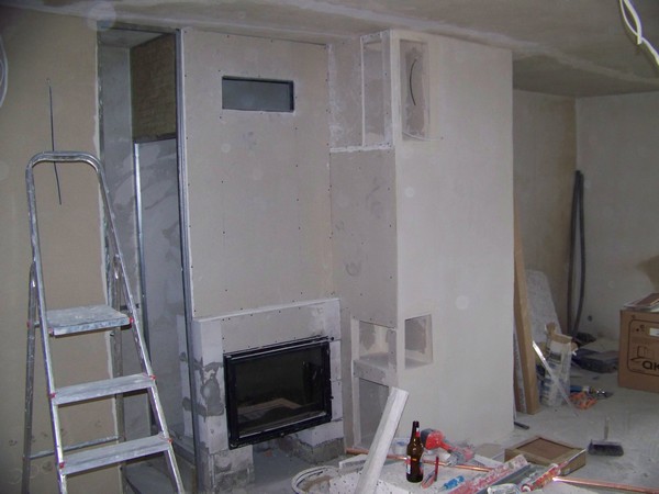 Montaż kominka z obudową z płyt gipsowo-kartonowych w trakcie prac wykończeniowych, widoczna drabina, narzędzia i materiały budowlane.