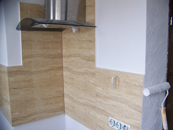 Fragment kuchni w trakcie remontu: ściana wyłożona płytkami imitującymi trawertyn, okap ze stali nierdzewnej i szkła, pomalowana na biało ściana z wystającymi kablami oraz wałek do malowania oparty...