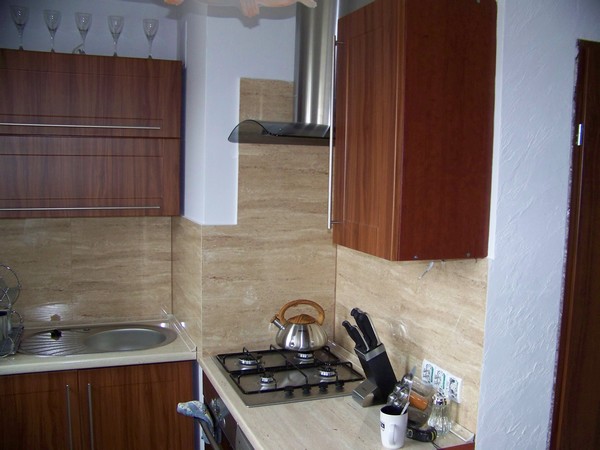 Wykończona kuchnia z brązowymi szafkami, beżowymi płytkami imitującymi marmur, stalowym okapem i płytą gazową z czajnikiem, widok na ścianę z gniazdkami.