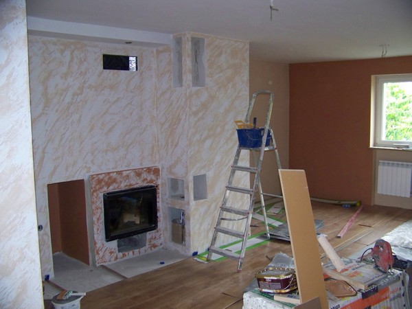 Wnętrze w trakcie remontu z widocznym kominkiem w zabudowie, ściany pokryte dekoracyjnym tynkiem w odcieniach beżu i brązu, drabina malarska z wiadrem, narzędzia malarskie i fragment podłogi z paneli.