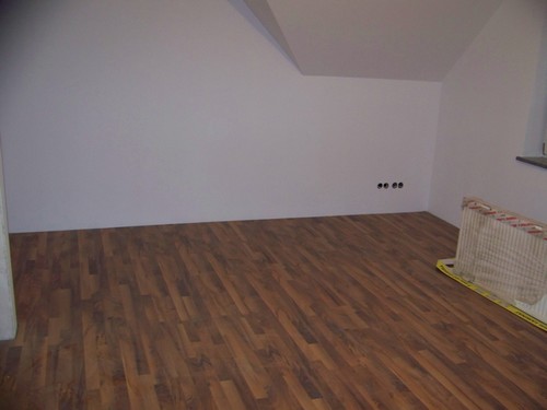 Pokój na poddaszu z brązową podłogą z paneli, białe ściany i widoczne puszki elektryczne bez osprzętu, w rogu oparty o ścianę grzejnik panelowy.