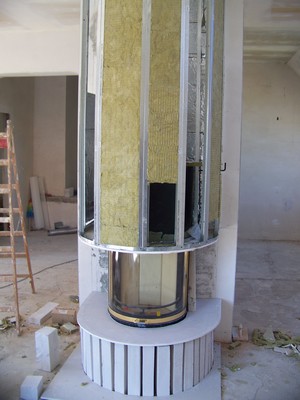 Wnętrze w trakcie budowy kominka z wkładem szklanym, widoczne materiały izolacyjne i metalowa konstrukcja szkieletowa, drabina w tle.