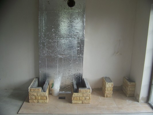 Wnętrze w trakcie budowy kominka z obudową z jasnej cegły, zabezpieczoną folią aluminiową ścianą i fragmentem podłogi z płytek.