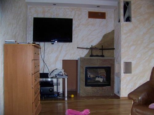 Salon z kominkiem obudowanym kamieniem, nad którym umieszczono dekoracyjny miecz samurajski, drewniana komoda z szufladami po lewej stronie, na ścianie telewizor LCD.