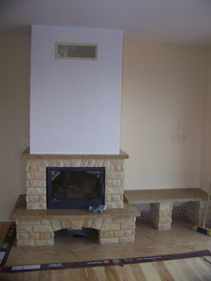 Nowoczesny kominek z obudową z jasnego kamienia, z prostokątnym wkładem kominkowym i szerokim blatem, umieszczony w surowym wnętrzu z jasnymi ścianami i częściowo wykończoną podłogą.