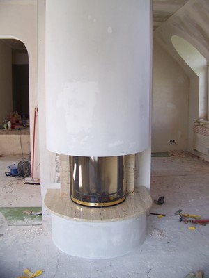 Nowoczesny kominek z okrągłą, białą obudową i metalowym wkładem, w trakcie instalacji w surowym wnętrzu z widocznymi śladami prac wykończeniowych.