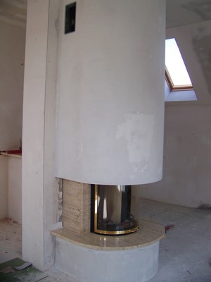 Wnętrze w trakcie wykończenia z owalnym kominkiem obudowanym jasnym kamieniem i gładzią, widoczne okno dachowe i surowy stan ścian.