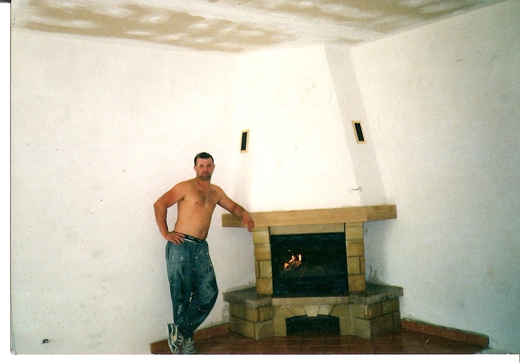 Mężczyzna bez koszulki, oparty o kamienny kominek z palącym się ogniem, w surowym wnętrzu z niedokończonym sufitem i wentylacją.